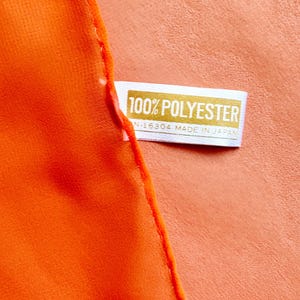 May include: A close-up shot of bright orange fabric with a label stating "100% POLYESTER" and "N-16304 MADE IN JAPAN". The label is white with gold text, and the fabric is a solid, vivid colour.