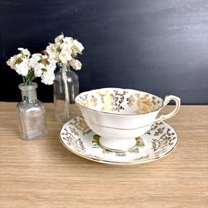 May include: White teacup and saucer set with gold filigree and trim. The teacup has a curved handle and sits on a matching saucer. The set is decorated with a floral and scroll design. A vintage tea set.