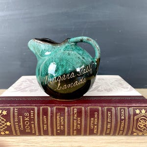 May include: A small green and brown ceramic pitcher with a handle. It has the words "Niagara Falls Canada" written on the side in gold lettering.