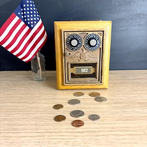 May include: A vintage-style mailbox with a wooden frame and a metal front, featuring two circular dials and the number 582. An American flag in a glass bottle stands to the left, with scattered coins in front of the mailbox.