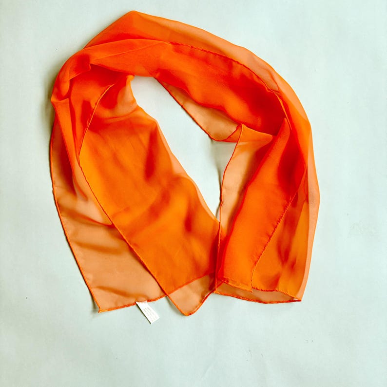 May include: Vibrant orange scarf crafted from a sheer, lightweight material. The scarf is arranged in soft folds, highlighting its delicate texture. A small label is attached. This accessory provides a burst of colour.