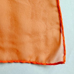 May include: Close-up of a square, orange fabric with a textured surface. The edges are finished with a bright orange, stitched border. The fabric has a soft, flowing appearance.