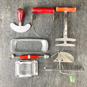 May include: A collection of vintage kitchen tools, including a red handled pastry cutter, a silver egg slicer, a silver pastry cutter, a silver cookie cutter, a silver melon baller, and a silver potato masher.