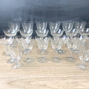 May include: A collection of clear, etched glass drinkware, including stemmed wine glasses, small bowls, and cordial glasses. The glasses have a textured pattern and are arranged on a light-colored wooden surface.
