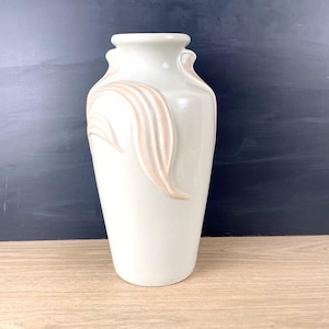 May include: A white ceramic vase with a textured design of pink leaves. The vase has a rounded body and a narrow neck.