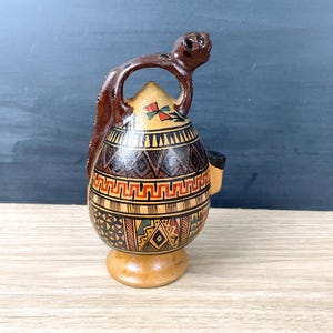 May include: A decorative ceramic pitcher with a brown handle and spout. The body is adorned with geometric patterns in shades of brown, tan, red, and green. The pitcher has a small base and a unique, sculpted handle. The overall design suggests a handcrafted, artisanal item.