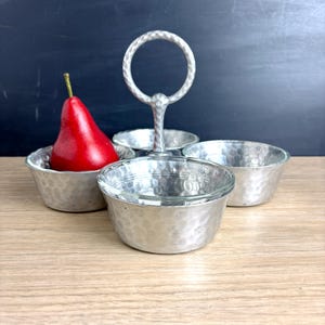 May include: A silver-colored serving dish with four hammered bowls and a handle. A red pear sits in one of the bowls. The dish is made of metal and has a textured surface. The handle has a circular design.