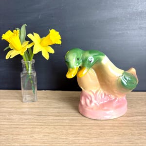 May include: A ceramic duck figurine with green heads and yellow beaks, resting on a pink base. The duck has tan and green wings. A small glass bottle with yellow daffodils is to the left of the duck.