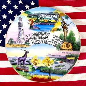 May include: A vintage ceramic plate with a colorful illustration of the Mohawk Trail in Massachusetts. The plate features a bridge, a mountain, a deer, and a Native American figure. The text "Mohawk Trail Massachusetts" is printed in black letters on the plate.