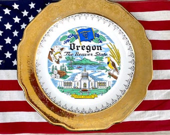 Puede incluir: Un plato decorativo con borde dorado y un diseño central con la bandera de Oregón, una vaca y el edificio del Capitolio Estatal. El plato está colocado sobre un fondo de la bandera estadounidense.
