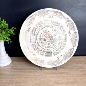 May include: A vintage white decorative plate with a gold floral border and calendar design. The plate features the year 1953, with a windmill illustration in the center. The calendar includes months from January to December.