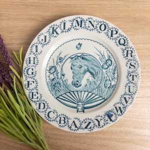 May include: A round ceramic plate featuring a dark blue transferware design on a cream background. The border displays the alphabet in capital letters. The center depicts a horse head surrounded by wheat stalks, a small butterfly, and a fan.