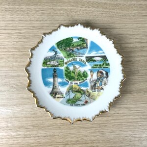 May include: A white decorative plate with a scalloped edge and gold trim. The plate features colorful illustrations of landmarks along the Mohawk Trail, including Elk Memorial and French King Bridge.