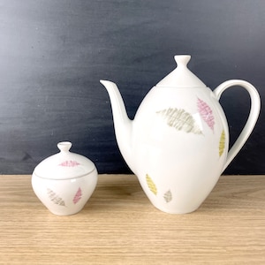 May include: A white ceramic teapot and sugar bowl with a leaf design in shades of pink, green, and gray. The teapot has a rounded body and a lid with a knob. The sugar bowl has a round body and a lid.