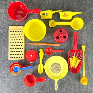 May include: A collection of yellow and red kitchenware items, including measuring cups, a colander, a drip pan, a grater, a strainer, and a set of skewers. The items are arranged on a gray surface.