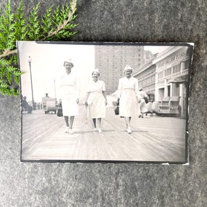 May include: Black and white photograph of three women wearing white dresses and hats walking on a boardwalk. The women are walking in a line, with the woman in the front leading the way. The boardwalk is made of wood and there are buildings in the background.