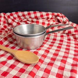 May include: A silver-colored metal colander nested inside a matching metal pot, both featuring long handles. A light-colored wooden spoon is positioned diagonally in the foreground. The items are set against a red and white checkered fabric.