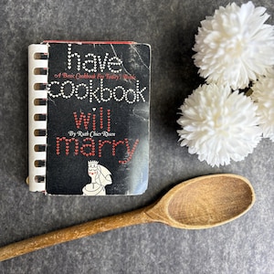 May include: A black and white cookbook with the title "Have Cookbook Will Marry" by Ruth Chier Rosen. The book is surrounded by white flowers and a wooden spoon.