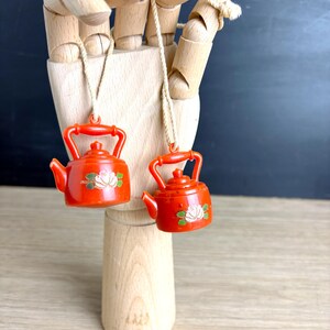 May include: Two small, red teapot pendants with white floral designs and matching handles hang from a wooden hand. The pendants are suspended by thin, beige cords. The background is a mix of wood and a dark surface.
