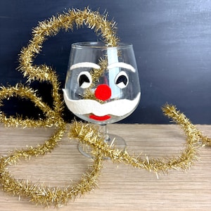 May include: A clear wine glass decorated with a Santa Claus face, featuring a white felt beard and mustache, black eyes, and a red nose and mouth. Gold tinsel is draped around the glass, creating a festive holiday scene.