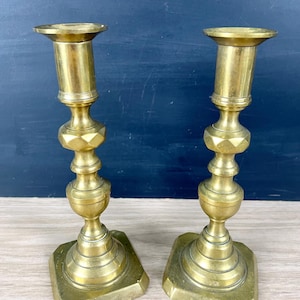 May include: A pair of matching brass candlesticks featuring a polished metallic finish, a wide flared top, a central faceted hexagonal section, and a tiered square base. These tall candle holders exhibit a classic vertical structure.