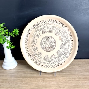 May include: Cream-colored vintage serving tray with a gold-colored calendar design. The tray features zodiac symbols and calendar dates. A decorative leaf border surrounds the calendar. The tray is displayed on a clear stand.