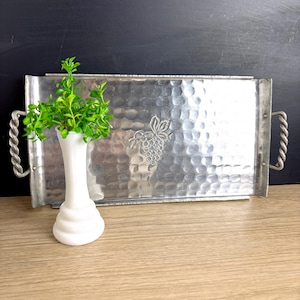 May include: A rectangular, hammered metal serving tray with twisted handles. A grape design is embossed on the tray's surface. A white vase with a green plant is on the tray. The tray is on a wooden surface.
