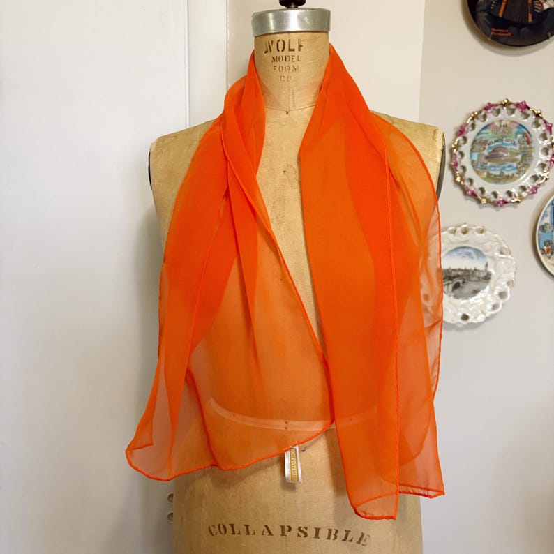 May include: An orange scarf on a vintage dress form. The scarf is made of a sheer, lightweight fabric. The dress form is a neutral beige colour, and the background includes decorative plates. The scarf's colour is a bright, eye-catching hue.