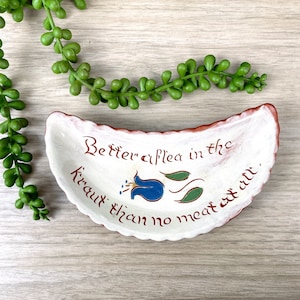 May include: A white ceramic dish shaped like a crescent moon with a red rim. The dish has a blue flower with green leaves and the text "Better a flea in the krau than no meat at all".