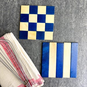 May include: Two wooden coasters with blue and natural wood designs. One coaster features a checkerboard pattern, while the other has vertical stripes. A red and white striped kitchen towel and a whisk are also in the image.