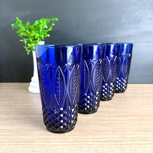 May include: Set of four cobalt blue drinking glasses. Each glass features a leaf and diamond pattern. The glasses are tall and cylindrical, perfect for water or other beverages. A white vase with greenery is in the background.