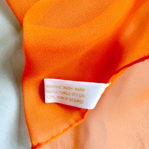 May include: Close-up of a bright orange scarf. The fabric is sheer and slightly translucent. A white tag with gold text reads "MACHINE WASH WARM, GENTLE TUMBLE DRY LOW, COOL IRON IF DESIRED."