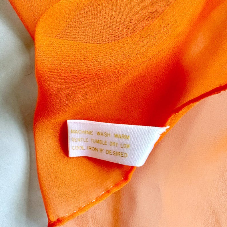 May include: Close-up of a bright orange scarf. The fabric is sheer and slightly translucent. A white tag with gold text reads "MACHINE WASH WARM, GENTLE TUMBLE DRY LOW, COOL IRON IF DESIRED."
