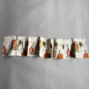 May include: A gathered fabric window valance featuring a repeating print of orange and red decorative vases and potted plants on a light beige background with a subtle white brick pattern. The valance has a rod pocket top for hanging.