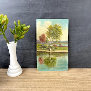 May include: A rectangular painting depicting a landscape with green and red trees along a calm body of water, reflecting the foliage. Beside the painting stands a white, smooth, multi-tiered vase containing a green succulent plant.