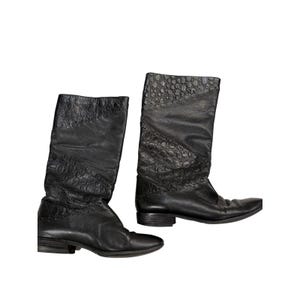 May include: A pair of black mid-calf boots featuring a combination of smooth leather and textured, embossed panels with a reptilian-like pattern. The boots have a rounded toe, a low block heel, and a slouchy shaft design with visible stitching.