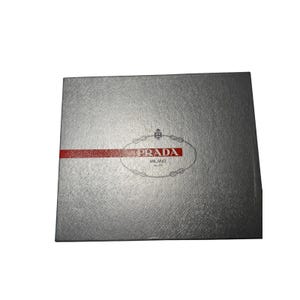 May include: A rectangular silver box with a textured surface featuring the Prada Milano logo printed in the center. A horizontal red stripe crosses the box, intersecting the logo which includes a small crest and the text Prada Milano dal 1913.