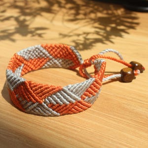May include: A handmade friendship bracelet in orange and gray, featuring a woven pattern. The bracelet has a sliding knot closure with wooden beads. The design is visible against a wooden surface.
