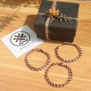 May include: Three braided bracelets with red, white, and black threads are displayed on a wooden surface. A black gift box tied with twine and decorated with a dried flower sits nearby. A white card with a logo and text is also present.