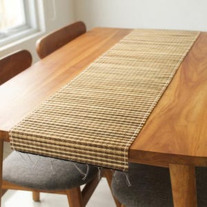 Pode incluir: Um caminho de mesa retangular tecido, em tons de castanho e bege, colocado sobre uma mesa de madeira. O caminho de mesa tem um padrão listrado e bordas com franjas, adicionando uma estética natural e rústica. A mesa é rodeada por cadeiras de madeira.