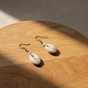 以下が含まれることがあります： シルバー色のフックと小さな白いビーズが付いた、クリーム色の貝殻のイヤリングのペア。 イヤリングは木製の表面に展示されており、自然光の影ができています。