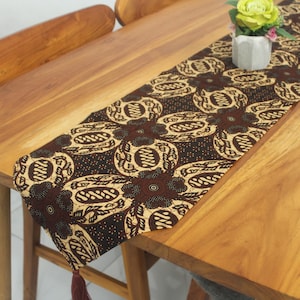 May include: A brown and beige batik table runner with a floral pattern. The runner is draped over a wooden table and features a repeating design of stylized flowers and circular motifs. A tassel hangs from one end.
