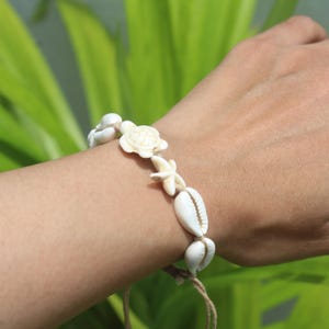 Könnte beinhalten: Ein weißes Muschelarmband mit einer Schildkröte, einem Seestern und verschiedenen Muschelformen, die auf einer naturfarbenen Kordel aufgereiht sind. Das Armband wird an einem Handgelenk vor unscharfem grünem Hintergrund gezeigt.