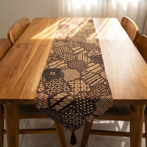 May include: A brown and tan patterned table runner with a tassel at the end, displayed on a wooden table. The runner features a repeating design of geometric shapes and patterns. The table is surrounded by wooden chairs.