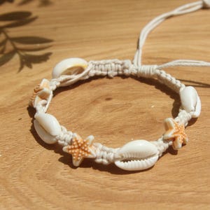Op de afbeelding: Een macramé armband met witte schelpen en oranje zeesterren. De armband is gemaakt met een crèmekleurig koord en is verstelbaar. Een accessoire met strandthema.