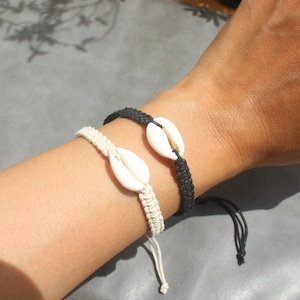 Puede incluir: Dos pulseras de macramé, una negra y otra crema, cada una con una gran concha blanca. Las pulseras están atadas con cordones ajustables. La imagen muestra las pulseras en una muñeca, mostrando un estilo playero y bohemio.