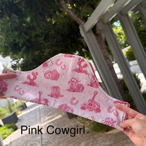 May include: A pink face mask with a repeating pattern of cowboy hats, cacti, guitars, and horseshoes. The text "Pink Cowgirl" is printed at the bottom of the image. The mask is held up by a person's hands.