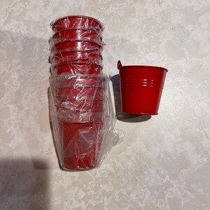 May include: A stack of small, red metal buckets wrapped in clear plastic, next to a single red bucket. The buckets have a classic bucket shape with a small handle on the side. The buckets are on a white and gray marble-patterned surface.
