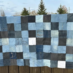 Könnte beinhalten: Eine Patchwork-Decke aus Jeansstoffquadraten in verschiedenen Blau- und Schwarztönen, mit einigen weißen Quadraten. Die Decke hängt im Freien an einem Holzzaun, mit Himmel und Bäumen im Hintergrund. Die Decke ist handgefertigt.