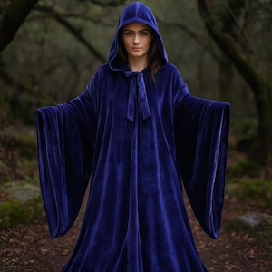 May include: A long, hooded cloak in a deep blue velvet fabric. The cloak features wide, flowing sleeves and a tie closure at the neck. The person is standing in a forest setting.